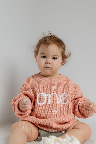 First birthday cake smash photo with a baby in pink sweater and a white birthday cake