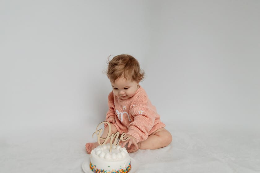 First birthday cake smash photo with a baby in pink sweater and a white birthday cake