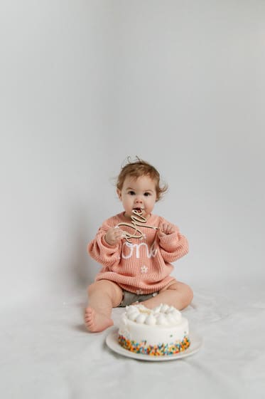 First birthday cake smash photo with a baby in pink sweater and a white birthday cake