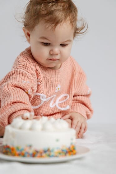 First birthday cake smash photo with a baby in pink sweater and a white birthday cake